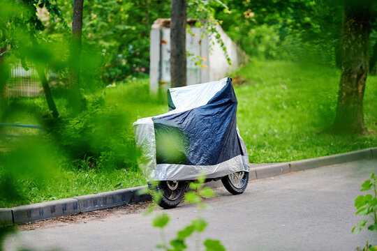 Motorcycle under protective blue silver cover on road next to green park area in soft natural daylight. Covered motorcycle on paved path with dense trees grass and out of focus leaves in foreground