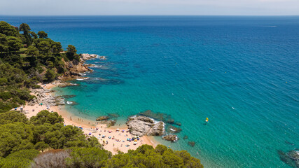 Obraz premium Aerial view of Cala Sa Boadella in Lloret de Mar, featuring a sandy beach, turquoise water, rocky coastline and lush Mediterranean vegetation.