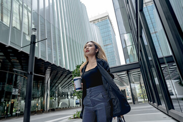 Fototapeta premium Asian businesswoman with wavy blonde hair looks upward while walking beneath towering glass skyscrapers, gray laptop bag over her shoulder, typical of a contemporary office lifestyle.