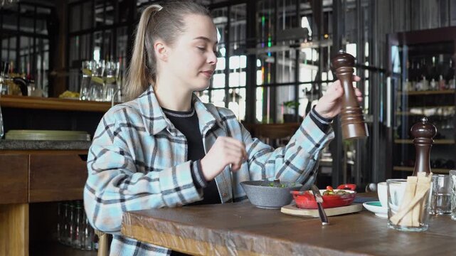 young girl salts dish at table in restaurant and smiles, woman adds pepper to salad in cafe