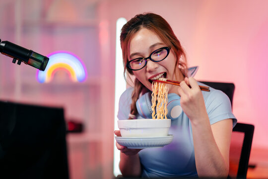 Young female streamer enjoying ramen noodles during gaming broadcast session at home studio with vibrant neon lighting