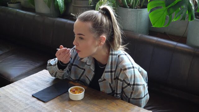 young girl eats small dessert with spoon in cafe and smiles, woman tries creme brulee in restaurant and shows happy emotions