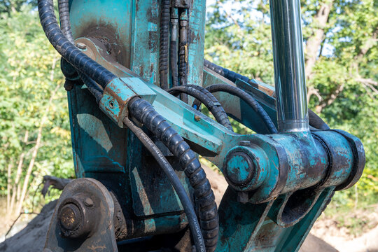 Close-up view of a heavy excavator arm with black hydraulic hoses, metal joints, and a chrome piston cylinder, working at a construction site in a lush green forest environment under bright daylight.