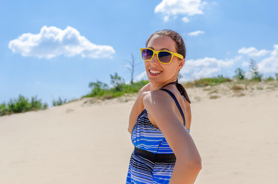 A smiling young woman wearing yellow sunglasses stands on a sandy dune under a bright blue sky.