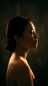 Serene profile portrait of an Asian woman with closed eyes in dramatic low light, suited for wellness, beauty, and meditation campaigns.