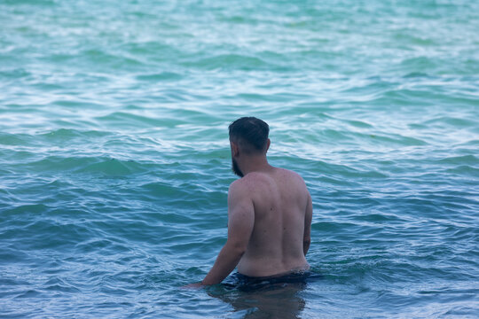 Rear view of a man standing waist-deep in clear turquoise ocean water, enjoying a calm and relaxing moment at the beach.
