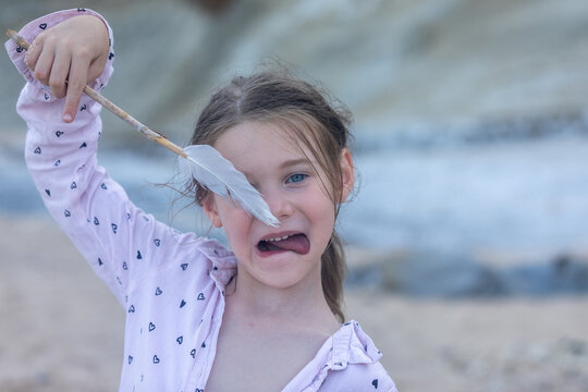 Smiling young girl holding a feather stick at the beach, making a funny face. Natural light, casual outdoor moment, carefree childhood.