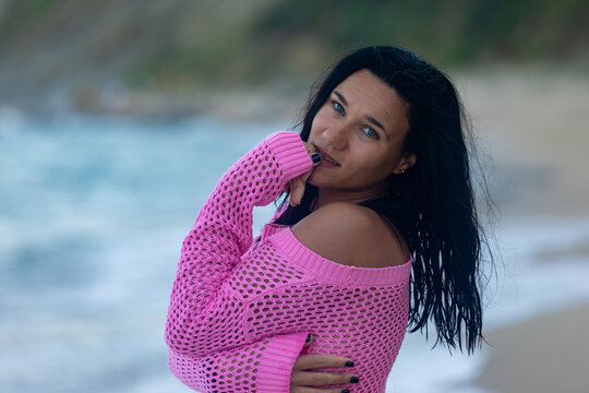 Confident woman with dark hair in a pink sweater poses on a sandy beach with waves and cliffs in the background.