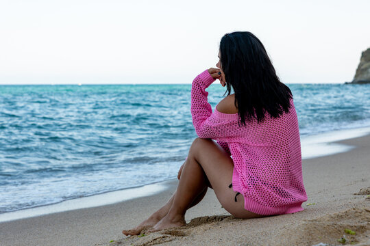 Woman in a pink sweater sitting alone on sandy beach, gazing at the ocean. Serene coastal scene with calm blue water and natural light.