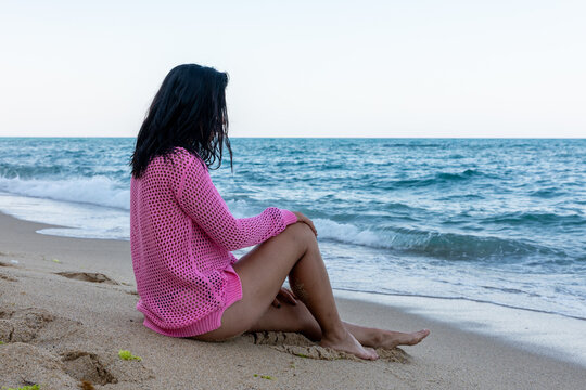 Woman in pink cover-up sitting on sandy beach, gazing at calm blue sea. Peaceful coastal scene perfect for vacation, wellness, or lifestyle themes.