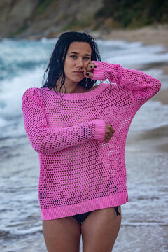 Young woman in a vibrant pink sweater and black bikini standing on a scenic beach with ocean waves and rocky cliffs in the background.