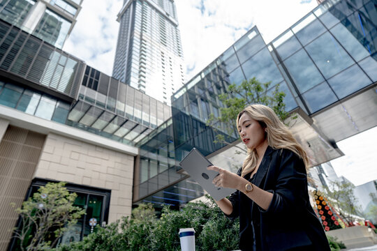 Asian businesswoman walks past a modern office entrance lined with greenery, eyes fixed downward on her tablet screen while a takeaway coffee cup rests on a planter beside the path.