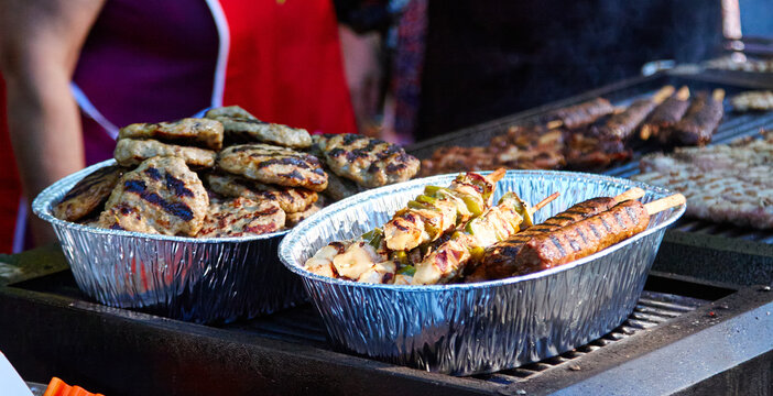 Grilled Bulgarian meat kufte and kebapche served in aluminum trays at a street food stall with smoke rising from the grill