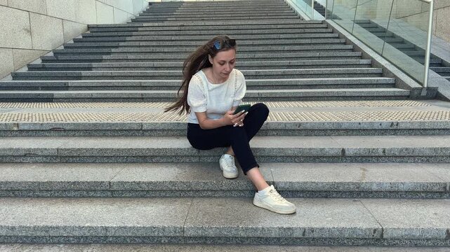 Young woman sitting on stone steps and looking at her smartphone, urban lifestyle, mobile technology, casual outdoor break.