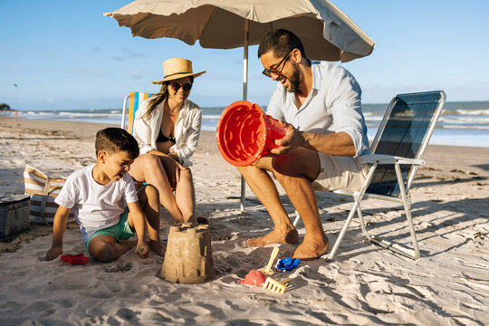 A happy family enjoys a sunny beach day as a father helps his son build a sandcastle while the mother relaxes under an umbrella, capturing playful summer moments and seaside bonding by the ocean.