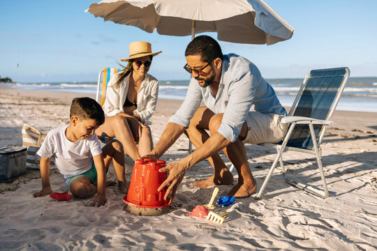 A happy family enjoys a sunny beach day as a father helps his son build a sandcastle while the mother relaxes under an umbrella, capturing playful summer moments and seaside bonding by the ocean.