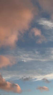 Soft peach and pink clouds drifting across a blue sunset sky background. Warm atmospheric light and peaceful summer weather, dreamy seasonal nature backdrop