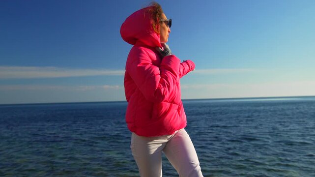 Woman sea jacket middle aged female in a bright pink puffer coat gesturing towards the ocean while enjoying a sunny day outdoors