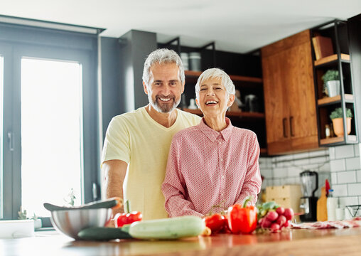 Portrait of happy senior mid aged mature couple prepering meal with fresh vegatebles in kitchen at hpme