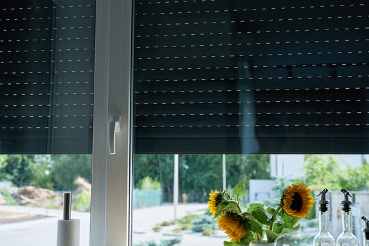 Partially closed roller shutters reduce daylight through a kitchen window, with sunflowers and glass bottles on the counter