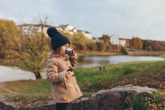 Cute little girl in beige coat and blue knit hat drinking from small metal mug holding cookie in autumn park. Childhood, autumn vibes, simple pleasures, seasonal lifestyle.