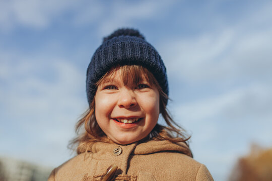 Close-up portrait of  joyful 4-year-old girl wearing blue beanie and beige coat, smiling on sunny autumn day. Childhood innocence, pure happiness, and joy of outdoor activities.