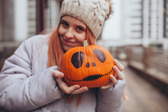 Young girl holding hand-painted Halloween pumpkin outdoors in autumn. Festive and cozy spirit of autumn season, Halloween traditions and joy of preparing for fall holidays.