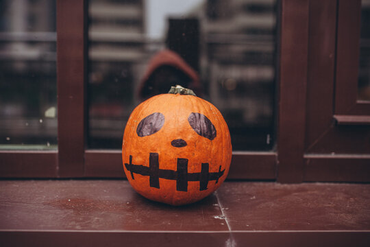 Small, hand-painted orange pumpkin with a scary face decoration sitting on windowsill. DIY spirit of Halloween preparations. Urban city decor.
