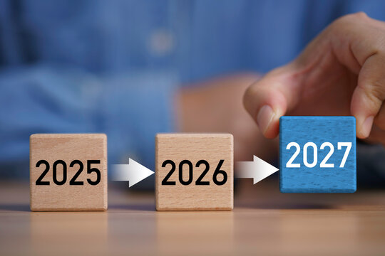 Hand placing 2027 wooden block after 2025 and 2026, symbolizing New Year planning, future goals, business growth, timeline progress strategy, transition, success and forward development.