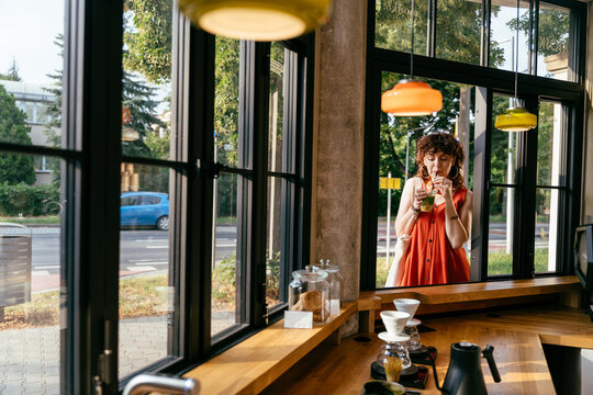 Stylish mature woman drinking iced matcha by cafe window in contemporary urban coffee shop