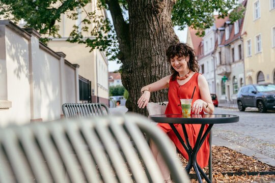 Cheerful mature woman enjoying iced matcha at outdoor cafe table on European city street