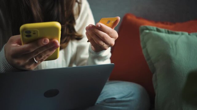 Anonymous young woman shopping online from her sofa with a credit card, shown in close-up while completing a digital purchase from home.
