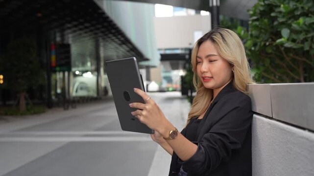 Asian businesswoman leans against a stone wall using her tablet, smiling and tilting her head back while standing on an empty city street in the Bangkok business district at dusk.