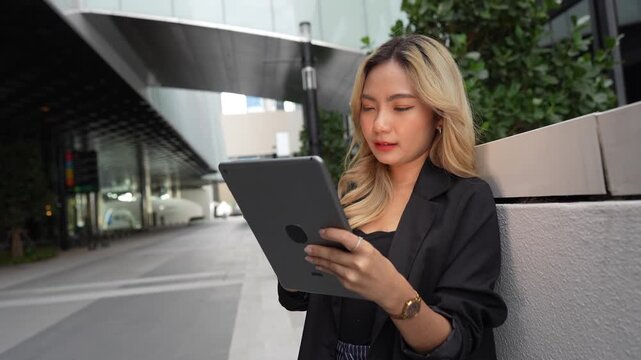 Asian businesswoman stands outdoors holding a stylus to her tablet, looking up thoughtfully before writing notes on the screen near a glass office building in the Bangkok business district.