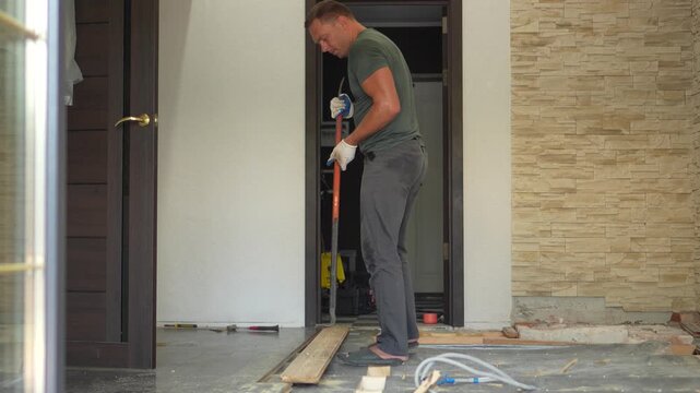 A cinematic medium shot of a strong man using a crowbar tool to pull up wooden floorboards during a house renovation.