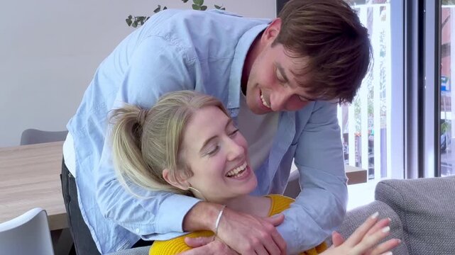 Cheerful caucasian couple laughing and talking affectionately while spending quality time together at home, with the smiling man embracing the woman from behind as she happily gestures