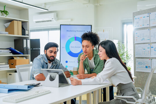 A hand reaches to point at a tablet screen amid folders and documents, colleagues leaning in to follow along a close, process-driven moment of collaborative problem-solving at work
