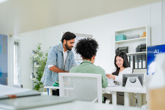 A man presents market share data on a large screen while explaining to two colleagues at a desk, one reviewing stock charts on a laptop a comprehensive scene of business analysis and team presentation