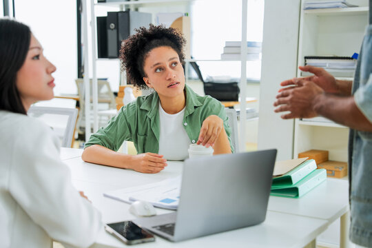 A woman listens attentively while a colleague gestures explanatorily off to the side, laptop and documents on the desk before her a candid moment of focused listening during a business meeting.