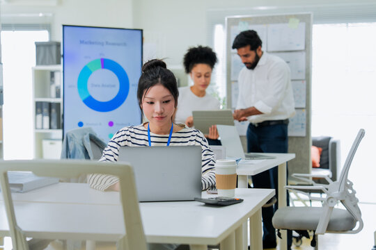 A focused employee works on her laptop in the foreground while colleagues collaborate at a desk behind her, capturing the layered activity of a busy, modern open-plan office in full swing.