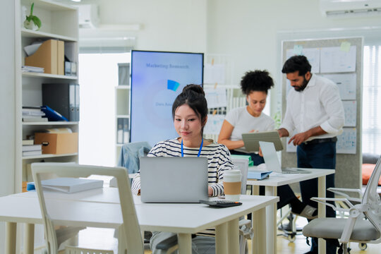 A focused employee works on her laptop in the foreground while colleagues collaborate at a desk behind her, capturing the layered activity of a busy, modern open-plan office in full swing.