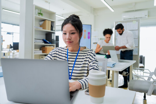 A focused employee works on her laptop in the foreground while colleagues collaborate at a desk behind her, capturing the layered activity of a busy, modern open-plan office in full swing.