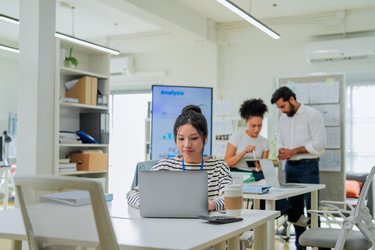 A focused employee works on her laptop in the foreground while colleagues collaborate at a desk behind her, capturing the layered activity of a busy, modern open-plan office in full swing.
