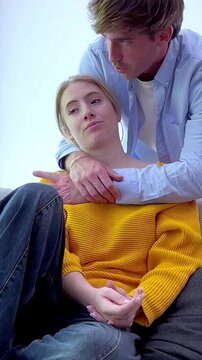 Young couple having a serious conversation while sitting on a sofa, with the man embracing the woman from behind as they discuss their problems and try to find a solution together