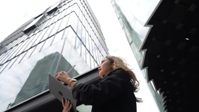 Asian businesswoman holds her tablet close while turning her head beneath soaring glass skyscrapers, framed in a dramatic extreme low angle shot within the modern Bangkok business district.