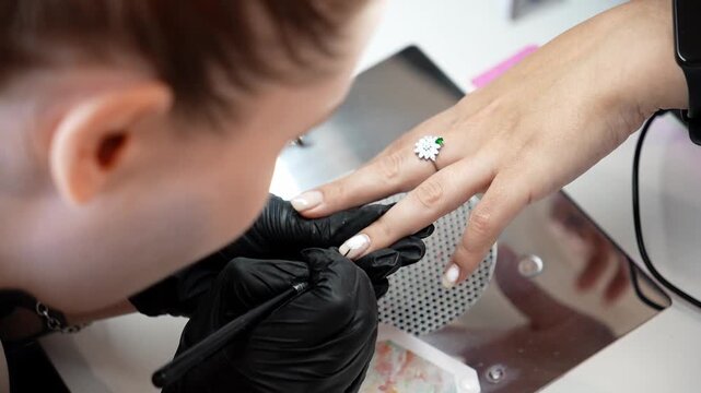 Nail technician paints a small white circle design on a clients fingernail using a fine brush. Creative nail art, precision work and professional manicure concept