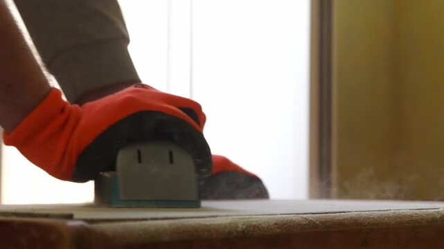 A close-up of a hand wearing an orange glove using a power sander on a wooden surface. The focus is on the tool and the sanding process.