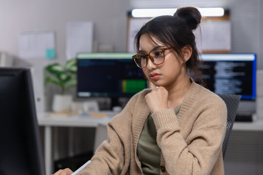 Woman office computer glasses desk work monitor sweater thoughtful programmer, young woman wearing glasses working desk modern office, looking thoughtful while using computer monitor and writing