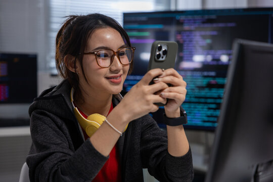 Woman programmer smartphone office computer code monitor glasses headphone technology, Asian woman programmer using smartphone office with computer monitor, code screen, glasses, yellow headphone