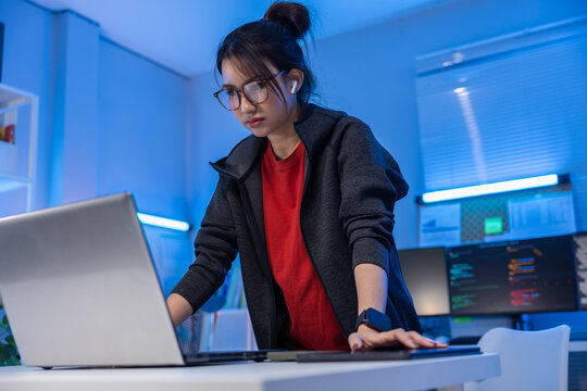 Asian woman laptop office glasses earbud red shirt dark hoodie focused expression blue light standing at desk while working in modern technology workspace